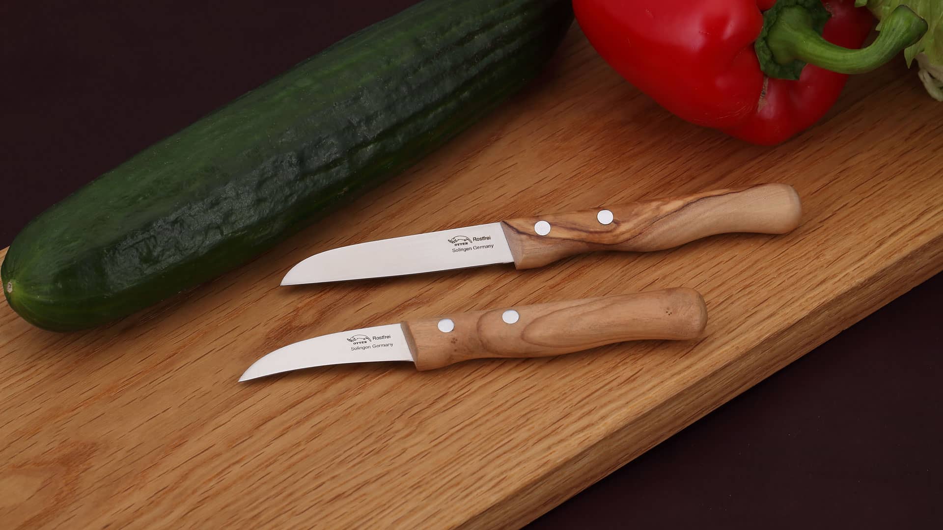 Schälmesser Zwei Küchenmesser mit Holzgriff auf einem Holzbrett neben einer Gurke und einer roten Paprika.